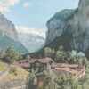 A small village with red-roofed buildings and a church sits in a green valley surrounded by steep cliffs and mountains under a blue sky. Nature and landscape poster print available in A1 to A4 sizes.