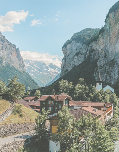 A small village with red-roofed buildings and a church sits in a green valley surrounded by steep cliffs and mountains under a blue sky. Nature and landscape poster print available in A1 to A4 sizes.