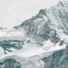 Snow-covered mountain peak with rocky, jagged ridges and patches of ice under a cloudy sky. Nature and landscape poster print available in A1 to A4 sizes.