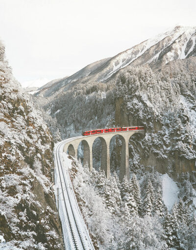 A red train crosses a high stone railway bridge near Tiefencastel, surrounded by snow-covered trees and mountains in Switzerland on a winter day. Nature and landscape poster print available in A1 to A4 sizes.