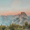 View of Half Dome rock formation in Yosemite National Park at sunset, with green shrubs in the foreground and pink clouds in the sky. Nature and landscape poster print available in A1 to A4 sizes.