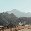 Rocky desert landscape with scattered shrubs in the foreground and mountains in the background under a clear sky. Dust rises from the ground near the rocks. Nature and landscape poster print available in A1 to A4 sizes.