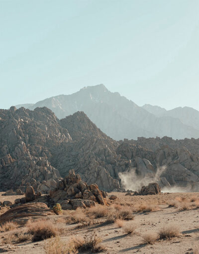 Rocky desert landscape with scattered shrubs in the foreground and mountains in the background under a clear sky. Dust rises from the ground near the rocks. Nature and landscape poster print available in A1 to A4 sizes.