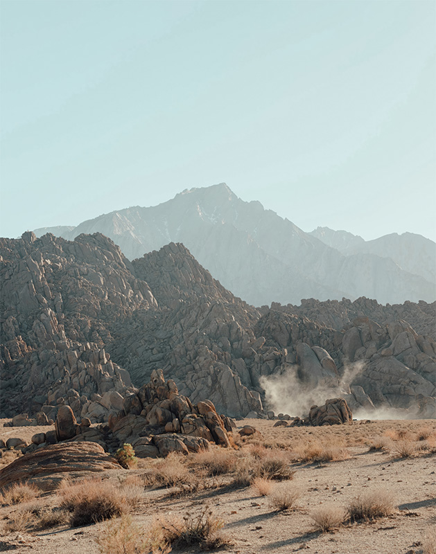 product-image Rocky desert landscape with scattered shrubs in the foreground and mountains in the background under a clear sky. Dust rises from the ground near the rocks. Nature and landscape poster print available in A1 to A4 sizes.