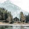 A wooden cabin sits by a lake, surrounded by pine trees, with a snow-capped mountain rising in the background under a cloudy sky. Nature and landscape poster print available in A1 to A4 sizes.