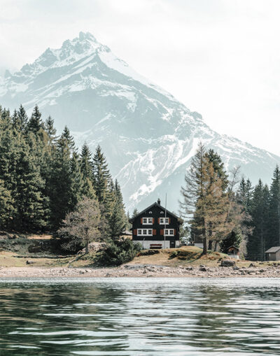 A wooden cabin sits by a lake, surrounded by pine trees, with a snow-capped mountain rising in the background under a cloudy sky. Nature and landscape poster print available in A1 to A4 sizes.