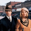 A man and woman in vintage 1930s attire stand side by side, each holding a gun, with classic cars and storefronts behind them. The text reads Bonnie and Clyde, referencing the iconic crime film from the 1967 movie. Movie poster print available in A1 to A4 sizes.