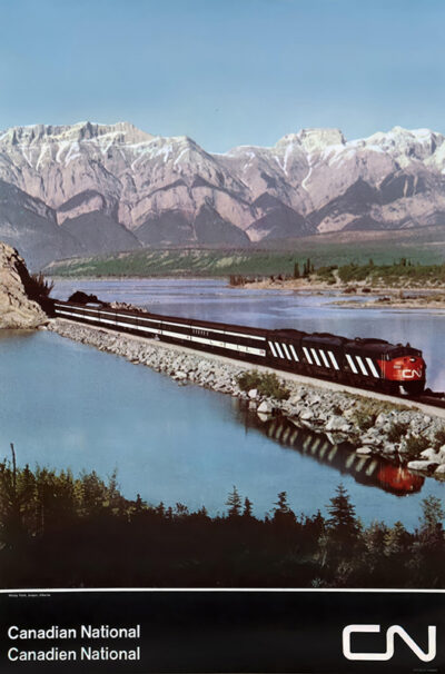 A Canadian National train travels along the lakeshore near Windy Point, with majestic mountains in the background under a clear blue sky in Jasper, Alberta. Poster print available in A1 to A4 sizes.